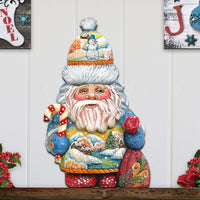 Decorative Christmas gnome Santa claus with colorful hat, scarf and candy cane on a wooden shelf against a white wall.