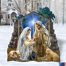 A large decorative Holy Night Christmas nativity scene sign displayed outdoors in the snow. The sign depicts Mary, Joseph, baby Jesus, a sheep under a starry blue sky framed by palm trees.