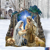 A large decorative Holy Night Christmas nativity scene sign displayed outdoors in the snow. The sign depicts Mary, Joseph, baby Jesus, a sheep under a starry blue sky framed by palm trees.