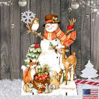 A festive Christmas scene featuring a storybook snowman with a striped orange scarf and brown hat, surrounded by woodland animals including a deer, owl, fox, squirrel, and duck. The scene includes hanging ornaments, a snowflake, string lights, and a rustic wood plank backdrop, with a small 'Made in USA' badge in the lower right corner.
