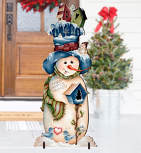 A decorative snowman yard sign with a tall blue hat, green scarf, and birdhouse details, displayed in front of a front door with a blurred Christmas tree in the background.