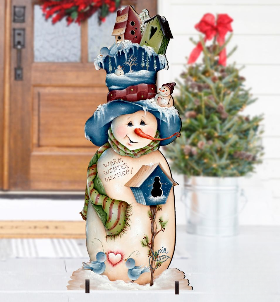 A decorative snowman yard sign with a tall blue hat, green scarf, and birdhouse details, displayed in front of a front door with a blurred Christmas tree in the background.