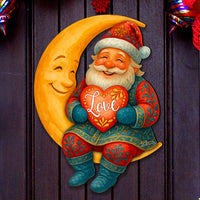 A festive holiday wall decor piece featuring a smiling Santa Claus sitting on a crescent moon and holding a red heart that says 'Love', displayed on a dark front door surrounded by red and green mesh garland and ornaments.