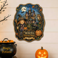 A decorative wall sign featuring an illustrated witch, haunted house, owl, pumpkin, and the text 'Witch's Potion Shop' displayed on a wood surface above a black cauldron and a glowing jack-o'-lantern against a white shiplap wall.