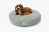 A small brown dog lies on a round, light sage green and white plaid designer cat and dog bed on a white background.