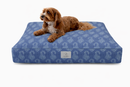 A small brown dog lies on a rectangular Designer Pet Bed with a Denim Blue Floral Paisley design against a clean white background. 