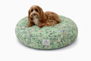 A small brown dog lies on a round green floral designer pet bed on a white background. The Chinoiserie cat and dog bed has a light green base, pink blossoms, bluebirds and leafy branches