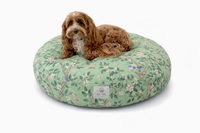 A small brown dog lies on a round green floral designer pet bed on a white background. The Chinoiserie cat and dog bed has a light green base, pink blossoms, bluebirds and leafy branches