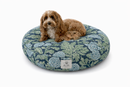 A small brown dog lies on a round designer pet bed with a blue and green floral botanical pattern in a Moody Maximalist style, on a white background.