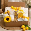A curated gift box set in a brown cardboard box, featuring a cream Knitted blanket with Tassel, Floral Bath Salts (16 oz.), soft bath puff, herbal tea (2 bags), honey sticks (2 sticks), ceramic tea caddy, cozy socks, Butterscotch Candies, 16 oz, and Glass tumbler with wood lid. The box is styled on a woven mat with lemons, a sunflower, and neutral decor in the background.