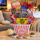 A movie lover's snack and candy gift basket arranged in a red-and-white striped popcorn bucket on a wooden table, with bowls of popcorn and candy in front. Visible items include Skittles, Sour Patch, M&M's, Airheads, Red Vines, Caramel Corn, and a 'Movie Night' sign in the background.