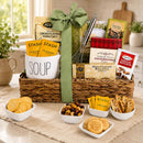 A woven gift basket filled with assorted snack, soup, cookies and food items, tied with a large green bow, displayed on a table with bowls of cookies, nuts, and dried fruit in a bright kitchen setting.