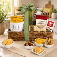 A woven gift basket filled with assorted snack, soup, cookies and food items, tied with a large green bow, displayed on a table with bowls of cookies, nuts, and dried fruit in a bright kitchen setting.