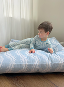 A young boy wearing a light blue and white gingham pajama set lies on a beachhouse blue and white patterned cat or dog bed in a room with sheer curtains and a wooden floor.
