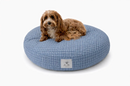 A small brown dog lies on a round blue-and-white plaid designer pet bed in a modern coastal style against a white background.