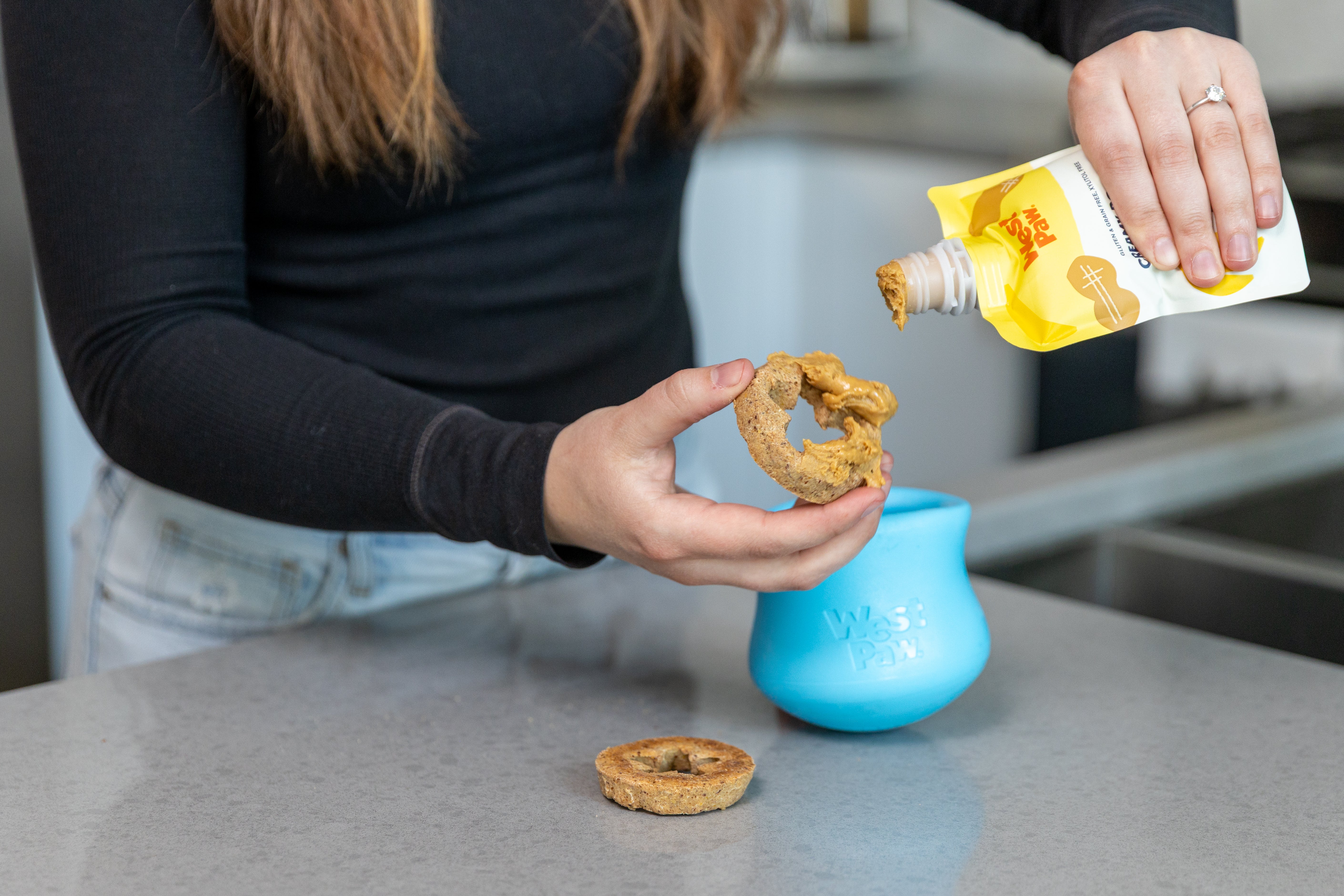 Person holding a Toppl dog biscuit next to a blue Toppl enrichment treat toy with a West Paw creamy squeezable treat puch on a kitchen counter.