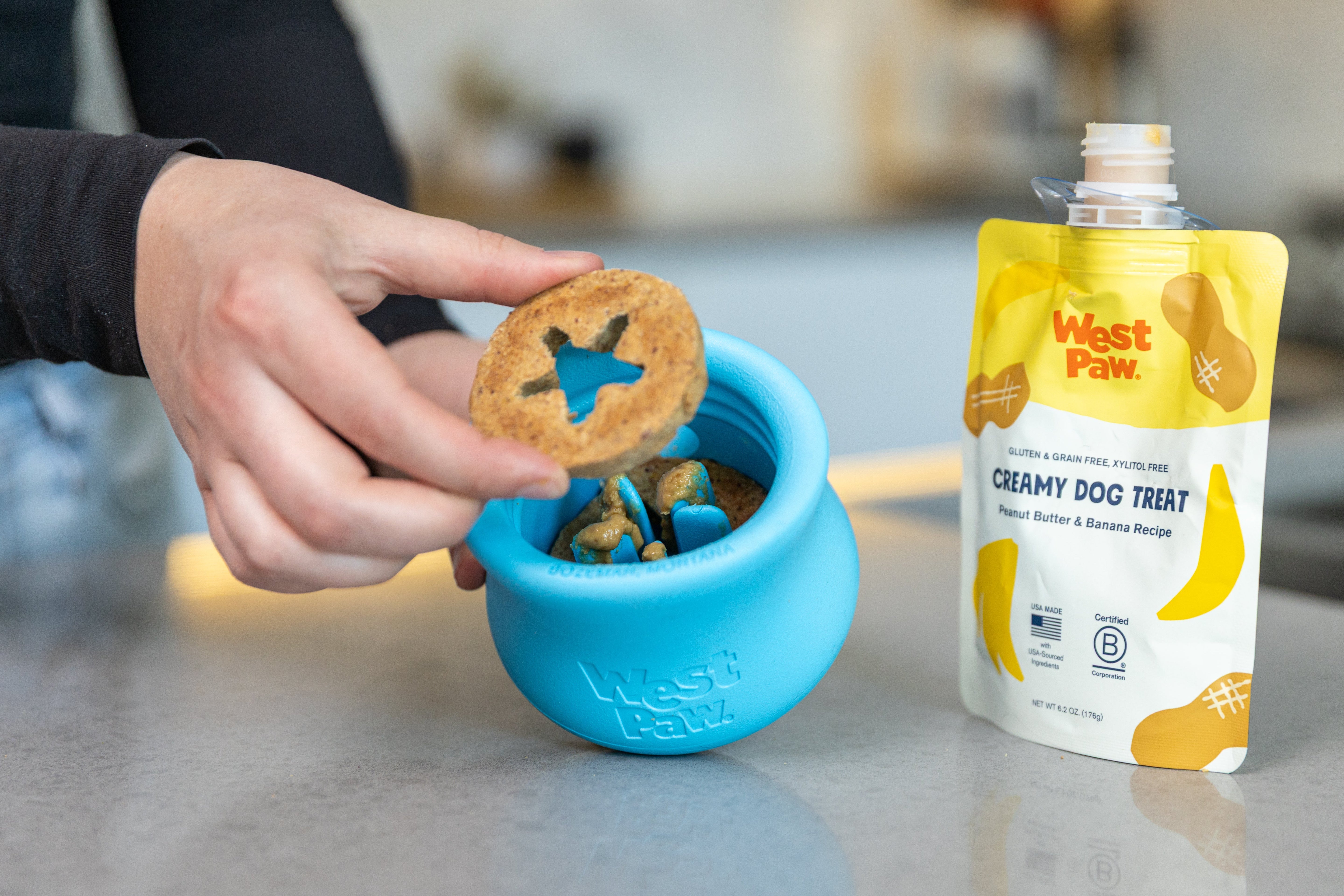Person holding a Toppl beef dog treat above a blue West Paw toppl toy with a West Paw peanut butter and banana creamy treat package in the background.