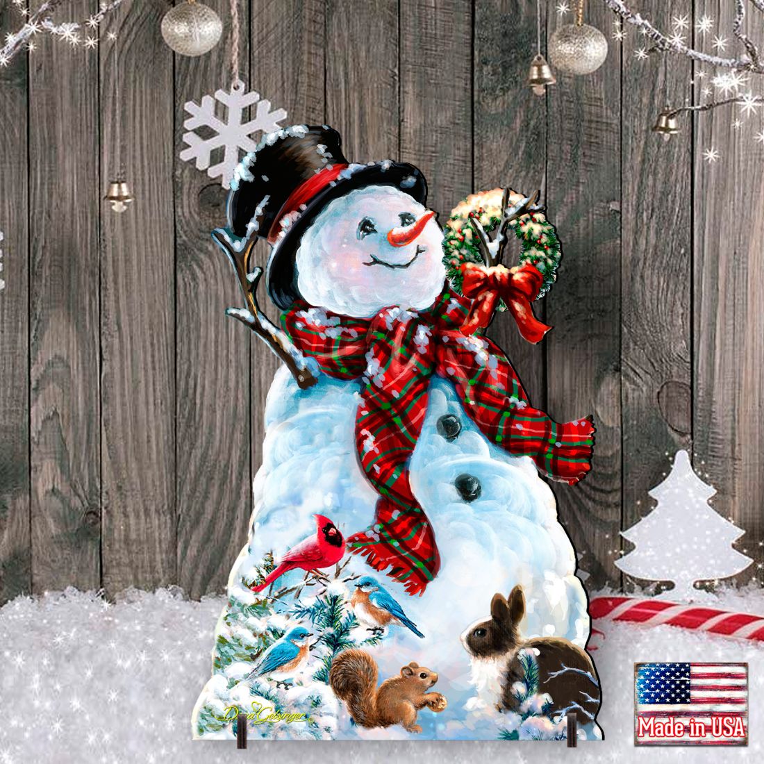 A snowman-shaped holiday wall decor piece with a black top hat, red plaid scarf, wreath, and winter animals printed at the base, shown against a rustic wood plank backdrop with hanging ornaments and string lights.