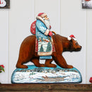 Decorative wall art of Santa Claus figure riding a bear on a wooden shelf.