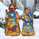 A decorative outdoor folk art Christmas nativity scene set featuring a standing wise man holding a scroll and a standing Mary holding baby Jesus, both with colorful printed robes and set in a snowy yard in front of a house.