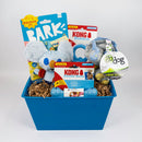 A pet toy gift basket in a blue plastic tub filled with dog toys and packaged treats. Visible items include a plush koala toy, blue balls, a Kong Binkie toy, a squeak ball, and a Go Dog chew guard, arranged in brown paper shred on a light gray background.