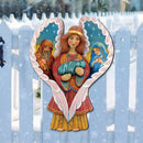 Decorative folk art painted nativity scene with an angel holding baby Jesus in front of a white picket fence on a snowy day.