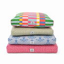 A stack of four square floor cushions in different patterns and colors, including a colorful patchwork design, a beige textured pattern, a light blue floral design, and a pink plaid design, on a white background.