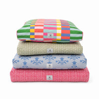A stack of four square floor cushions in different patterns and colors, including a colorful patchwork design, a beige textured pattern, a light blue floral design, and a pink plaid design, on a white background.