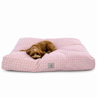 A brown dog lies on a blush pink barbiecore cat or dog bed on a clean white background. 