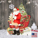 A festive Christmas-themed decorative scene featuring Santa Claus in a red suit sitting in a sleigh loaded with wrapped gifts reading a christmas list, beside a decorated Christmas tree. The background is a rustic dark wood plank wall with hanging ornaments and lights, and the foreground includes faux snow, a white snowflake, a small tree, and candy canes.