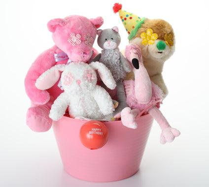 A pink plastic bucket filled with dog toys: plush stuffed animals, including a large pink bear, a white bunny, a gray cat, a tan bear wearing a party hat, and a pink flamingo figure. A red round tag on the front of the bucket reads 'HAPPY BIRTHDAY!'.