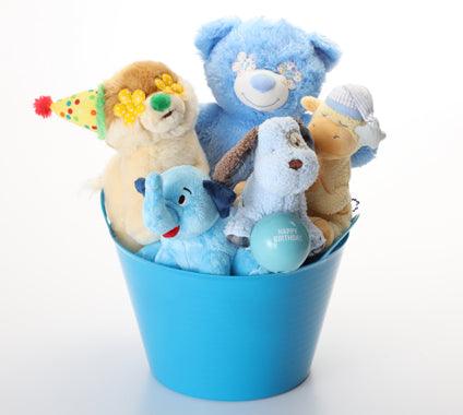 A blue bucket filled with plush stuffed animals, including a large blue bear, a tan bear with a party hat, a small blue elephant, and other soft toys, photographed on a white background.