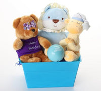 A plush toy gift basket in a bright blue box, featuring three stuffed animals: a brown bear wearing a purple 'Happy Birthday' shirt with purple flowers in its ears, a light blue lion with a white mane, and a beige giraffe wearing a blue and white hat. A light blue ball reading 'Happy Birthday' sits in front of them.