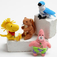 Four small plush cat toys arranged on white foam blocks against a light gray studio background: a yellow bird-like figure with a red neck scarf, a brown squirrel holding a nut, a blue-and-white bird perched on a block, and a pink star-shaped character wearing green and purple shorts.