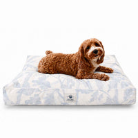 A small brown dog with curly fur lies on a rectangle dog bed with a modern light gray and white floral pattern, photographed against a clean white background.