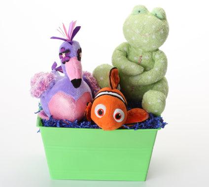 A plush toy gift basket featuring a green frog, a pink and purple flamingo, and an orange clownfish nestled in blue shredded paper inside a bright green basket, set against a plain white background.